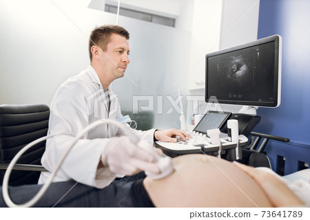 Side view portrait of professional focused pleasant Caucasian male doctor, doing ultrasound scan for pregnant woman in modern hospital. Focus on the doctor's face 73641789