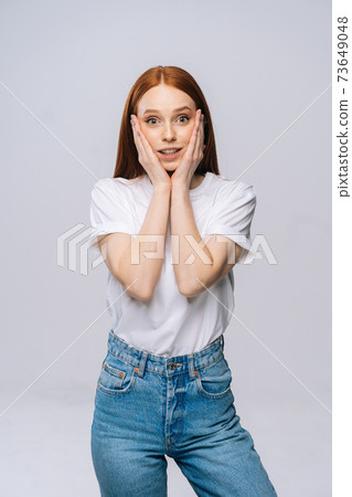 Amazed charming young woman wearing T-shirt and denim pants smiling with hands on cheek. 73649048