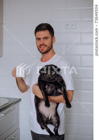 A man holds a pug dog in his arms, and drinks coffee A man holds a pug dog in his arms, and drinks coffee 73649504