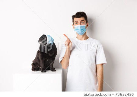 Covid-19, animals and quarantine concept. Dog owner and cute black pug wearing medical masks, man pointing and puppy staring at upper left corner, white background 73652486