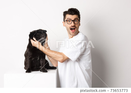 Happy young man showing you cute face of his pug. Dog owner loving his pet, standing over white background Happy young man showing you cute face of his pug. Dog owner loving his pet, standing over white background 73653773