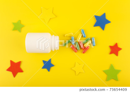 White medication bottle with oval multicolored pills spilled against yellow background decorated with stars 73654409