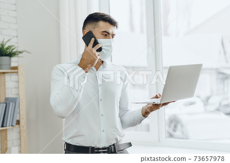 Handsome man businessman in medical mask using his laptop while talking on his cellphone 73657978