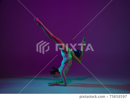 Young female athlete, rhythmic gymnastics artist on purple background with neon light. Beautiful girl practicing with equipment. Grace in performance. 73658597
