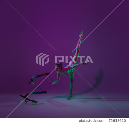 Young female athlete, rhythmic gymnastics artist on purple background with neon light. Beautiful girl practicing with equipment. Grace in performance. 73658610