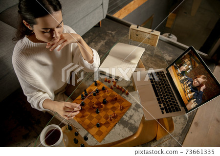 Young beautiful girl sitting at home and playing chess with friend online. Young beautiful girl sitting at home and playing chess with friend online. 73661335