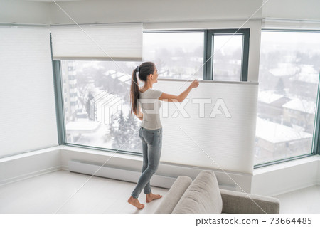 Woman opening home curtains in urban condo. Modern top down bottom up privacy cellular shades on apartment window keeping heat in winter with honeycomb blind curtain. Cordless pleated shades Woman opening home curtains in urban condo. Modern top down bottom up privacy cellular shades on apartment window keeping heat in winter with honeycomb blind curtain. Cordless pleated shades 73664485