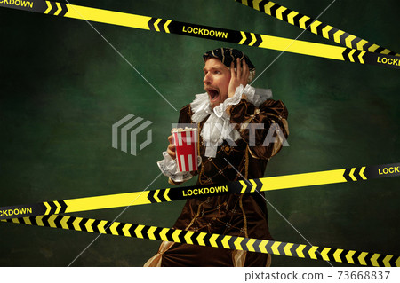 Medieval young man in old-fashioned costume with popcorn behing the limiting tapes with word Lockdown 73668837