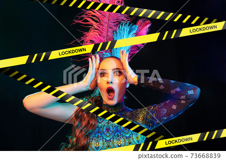Beautiful young woman in carnival, stylish masquerade costume with feathers behind limiting tapes with word Lockdown Beautiful young woman in carnival, stylish masquerade costume with feathers behind limiting tapes with word Lockdown 73668839
