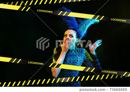 Beautiful young woman in carnival, stylish masquerade costume with feathers behind limiting tapes with word Lockdown Beautiful young woman in carnival, stylish masquerade costume with feathers behind limiting tapes with word Lockdown 73668889