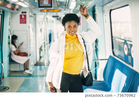 A smiling young African woman holds on to a handrail while standing on a subway train during her daily commute to work 73670962