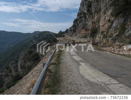 Narrow dangerous asphalt road at Genna Croce pass. at Supramonte Mountains with white limestone rocks, green hills, trees and mediterranean forest vegetation. Sardinia, Italy. Summer cloudy sky. Narrow dangerous asphalt road at Genna Croce pass. at Supramonte Mountains with white limestone rocks, green hills, trees and mediterranean forest vegetation. Sardinia, Italy. Summer cloudy sky. 73672236