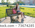 Woman in summer in a city park, resting after work, in hand a coffee tea cup. Rest and lunch after work, breakfast break. Green grass background and cars, scooter. 73674285