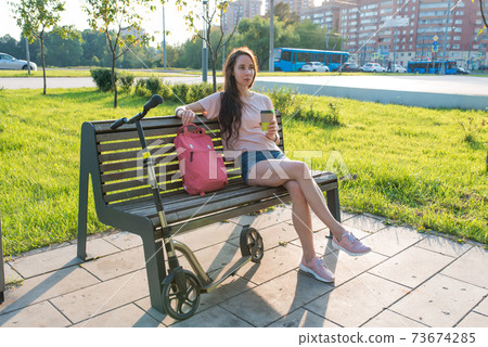 Woman in summer in a city park, resting after work, in hand a coffee tea cup. Rest and lunch after work, breakfast break. Green grass background and cars, scooter. Woman in summer in a city park, resting after work, in hand a coffee tea cup. Rest and lunch after work, breakfast break. Green grass background and cars, scooter. 73674285