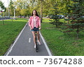 woman in summer in city rides a scooter on a bicycle path, in motion, background road trees green grass and spruce, pink jacket denim shorts. Free space for a copy of the text. 73674288