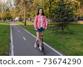 woman in summer in city rides a scooter on a bike path, background road trees green grass and spruce, pink jacket denim shorts. Free space for a copy of the text 73674290
