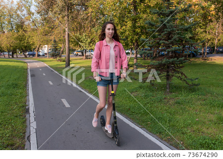 woman in summer in city rides a scooter on a bike path, background road trees green grass and spruce, pink jacket denim shorts. Free space for a copy of the text woman in summer in city rides a scooter on a bike path, background road trees green grass and spruce, pink jacket denim shorts. Free space for a copy of the text 73674290