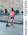 woman in summer in city goes to underground passage with a scooter, denim shorts and a pink jacket with a backpack. Compliance with road safety rules. Background road cars and buildings. 73674291