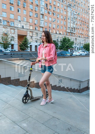 woman in summer in city goes to underground passage with a scooter, denim shorts and a pink jacket with a backpack. Compliance with road safety rules. Background road cars and buildings. woman in summer in city goes to underground passage with a scooter, denim shorts and a pink jacket with a backpack. Compliance with road safety rules. Background road cars and buildings. 73674291