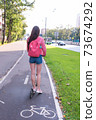 woman rides a scooter in city in summer, a view from back, denim shorts and a pink jacket with a backpack. The cycle path is wet after rain. Cars road background. 73674292