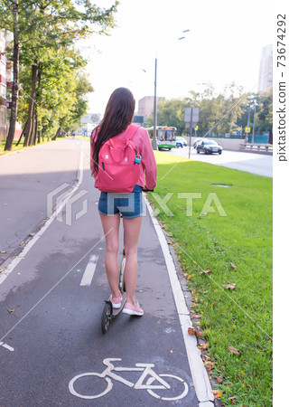 woman rides a scooter in city in summer, a view from back, denim shorts and a pink jacket with a backpack. The cycle path is wet after rain. Cars road background. woman rides a scooter in city in summer, a view from back, denim shorts and a pink jacket with a backpack. The cycle path is wet after rain. Cars road background. 73674292