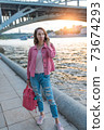 A woman in summer in city stands by river bank, making a phone call, dating and meeting on street, traveling and enjoying a weekend. Background bridge over the river. 73674293