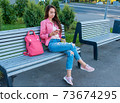 woman in city on bench in summer, reads writes message in chat, social networks on Internet, application smartphone. Casual wear, pink jacket, bag jeans. Relax of coffee tea after work. 73674295