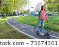Woman on a scooter, in summer in city rides in motion, free space for a copy of text. Pink jacket, backpack, jeans. Background green lawn, trees, bike path, road and cars. 73674301