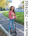 Woman on a scooter in summer in city. Stands and looks at route on phone. Compliance with road safety rules. Pink jacket and jeans. Route search and Internet navigation application. 73674302