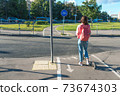 Girl on scooter summer in city. Stands with his back, crossroads, waiting for car to pass. Free space copy of text. Compliance with road safety rules. Pink jacket bag and jeans. Bicycle path sign. 73674303