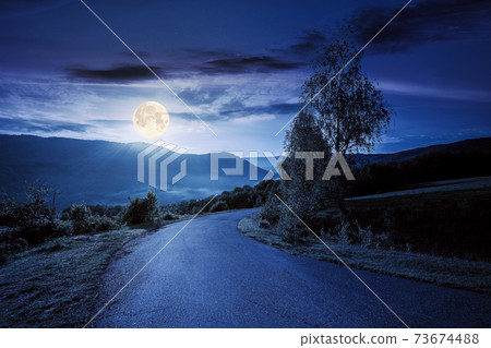 countryside road in mountains at night. beautiful view in to the distant foggy valley from the top of the pass. trees along the way in full moon light. wonderful rural landscape in summer 73674488