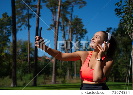 A happy woman with headphones listening to the music and making selfie outdoor 73674524