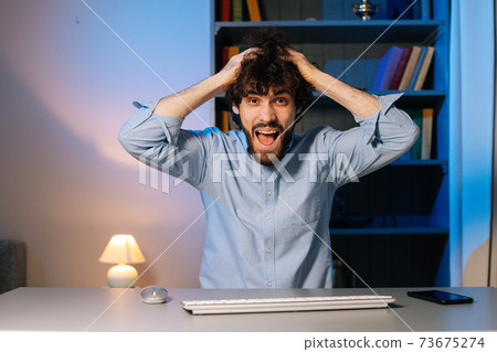 Front view of crazy young man pulling hair on head sitting at desk with computer at home office Front view of crazy young man pulling hair on head sitting at desk with computer at home office 73675274