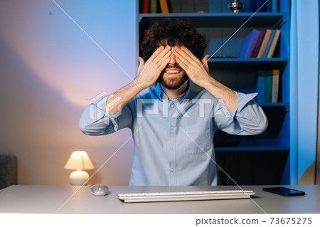 Front view of dissenting young man sitting at desk and covering face with hand. Front view of dissenting young man sitting at desk and covering face with hand. 73675275