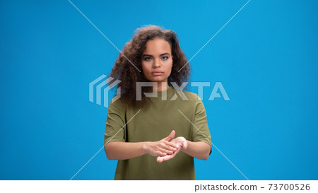 Using her hands to communicate pretty african american girl in reusable protective face mask. Curly haired young woman showing hello sign talking on camera isolated on blue background 73700526