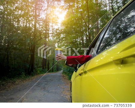Young woman holds out a takeaway paper cup with hot drink through the driver's seat window. Focus on paper cup Young woman holds out a takeaway paper cup with hot drink through the driver's seat window. Focus on paper cup 73705339