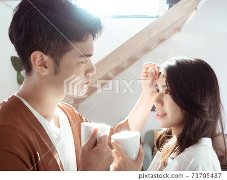 Young asian woman and man are enjoying spending time together at home with cup of coffee in hands. Young asian woman and man are enjoying spending time together at home with cup of coffee in hands. 73705487