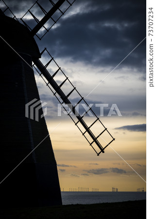 Traditional windmill silhouetted against modern offshore wind farm 73707234