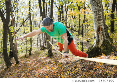 A bearded man in age balances while sitting on a taut slackline in the autumn forest. Outdoor Leisure A bearded man in age balances while sitting on a taut slackline in the autumn forest. Outdoor Leisure 73707499