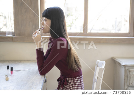 A woman sitting on a chair and applying lipstick by the window 73708536
