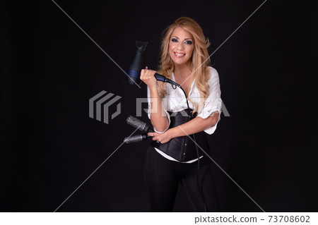 Blonde hairdresser holding a hairdryer and hair brushes while smiling on a black background. 73708602