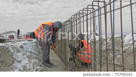 Engineers work with metal constructions while building a house. Builder fixes armature while working on construction site. Workers at joining metallic iron bars structure for reinforcement wall Engineers work with metal constructions while building a house. Builder fixes armature while working on construction site. Workers at joining metallic iron bars structure for reinforcement wall 73709191