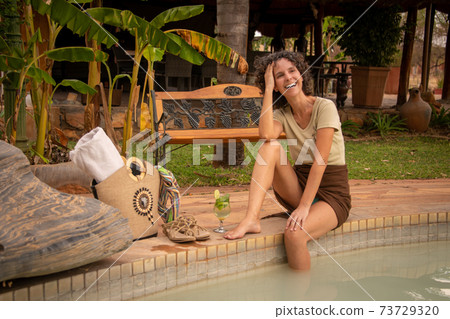 Laughing brunette sitting by pool with bag 73729320