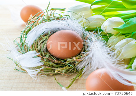 Basket easter decoration: natural colour eggs in basket with spring tulips, white feathers on wooden table background. Congratulatory easter design. 73730645