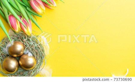 Easter Golden eggs in basket with spring tulips, white feathers on pastel yellow background in Happy Easter decoration. Spring holiday flat lay concept. 73730690