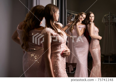 Two young beautiful girls wearing full-length pink violet or purple chiffon mermaid dresses or prom ball gowns decorated with sparkles and sequins. Models in front of mirror in a fitting room. Two young beautiful girls wearing full-length pink violet or purple chiffon mermaid dresses or prom ball gowns decorated with sparkles and sequins. Models in front of mirror in a fitting room. 73741148