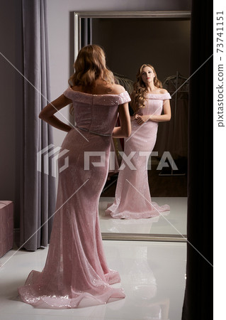 Young beautiful blonde girl wearing a full-length pink violet or purple chiffon mermaid dress or prom ball gown decorated with sparkles and sequins. Model in front of mirror in a fitting room. Young beautiful blonde girl wearing a full-length pink violet or purple chiffon mermaid dress or prom ball gown decorated with sparkles and sequins. Model in front of mirror in a fitting room. 73741151