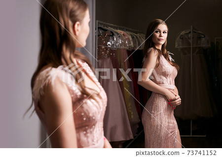 Young beautiful brunette girl wearing a full-length pale pink fitting chiffon prom ball slit gown decorated with sparkles and sequins. Model in front of mirror in a fitting room. Young beautiful brunette girl wearing a full-length pale pink fitting chiffon prom ball slit gown decorated with sparkles and sequins. Model in front of mirror in a fitting room. 73741152