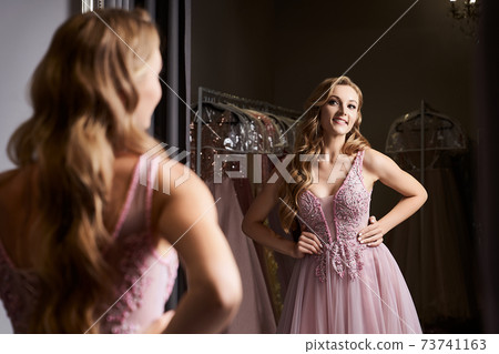 Young beautiful blonde girl wearing a full-length pink violet or purple chiffon prom ball gown decorated with sparkles and sequins. Model in front of mirror in a fitting room at dress hire service. Young beautiful blonde girl wearing a full-length pink violet or purple chiffon prom ball gown decorated with sparkles and sequins. Model in front of mirror in a fitting room at dress hire service. 73741163
