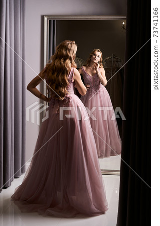 Young beautiful blonde girl wearing a full-length pink violet or purple chiffon prom ball gown decorated with sparkles and sequins. Model in front of mirror in a fitting room at dress hire service. Young beautiful blonde girl wearing a full-length pink violet or purple chiffon prom ball gown decorated with sparkles and sequins. Model in front of mirror in a fitting room at dress hire service. 73741166